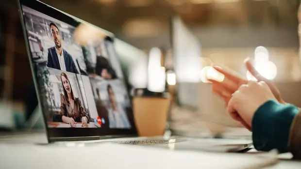 Hände bedienen ein Smartphone neben einem Laptop mit laufender Videokonferenz.